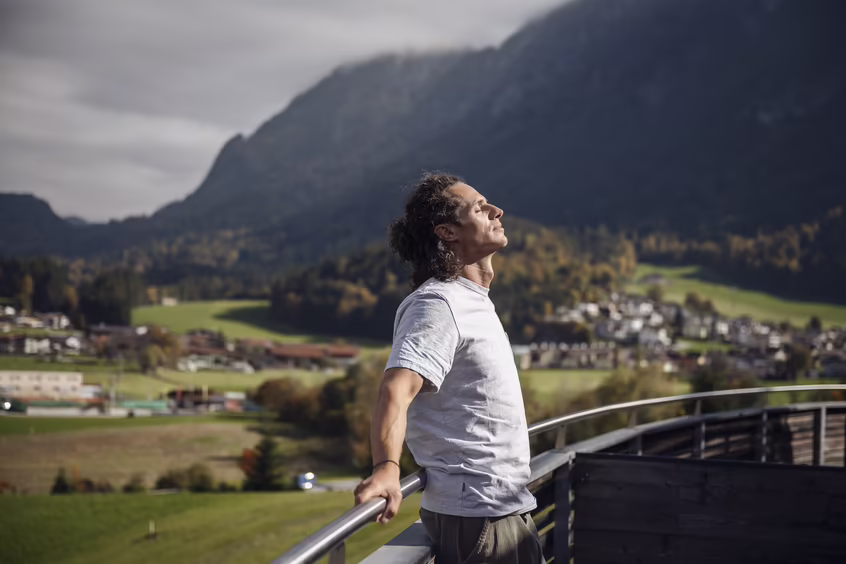 DAS SIEBEN - sieben Sinne - hören ©alexgretter Mann atmet auf einer Terrasse mit weitem Blick auf Berge und Landschaft im DAS SIEBEN durch